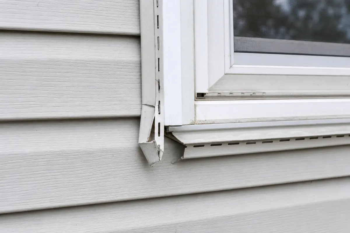 Damaged white vinyl J-channel cracked loose beside a window on gray siding
